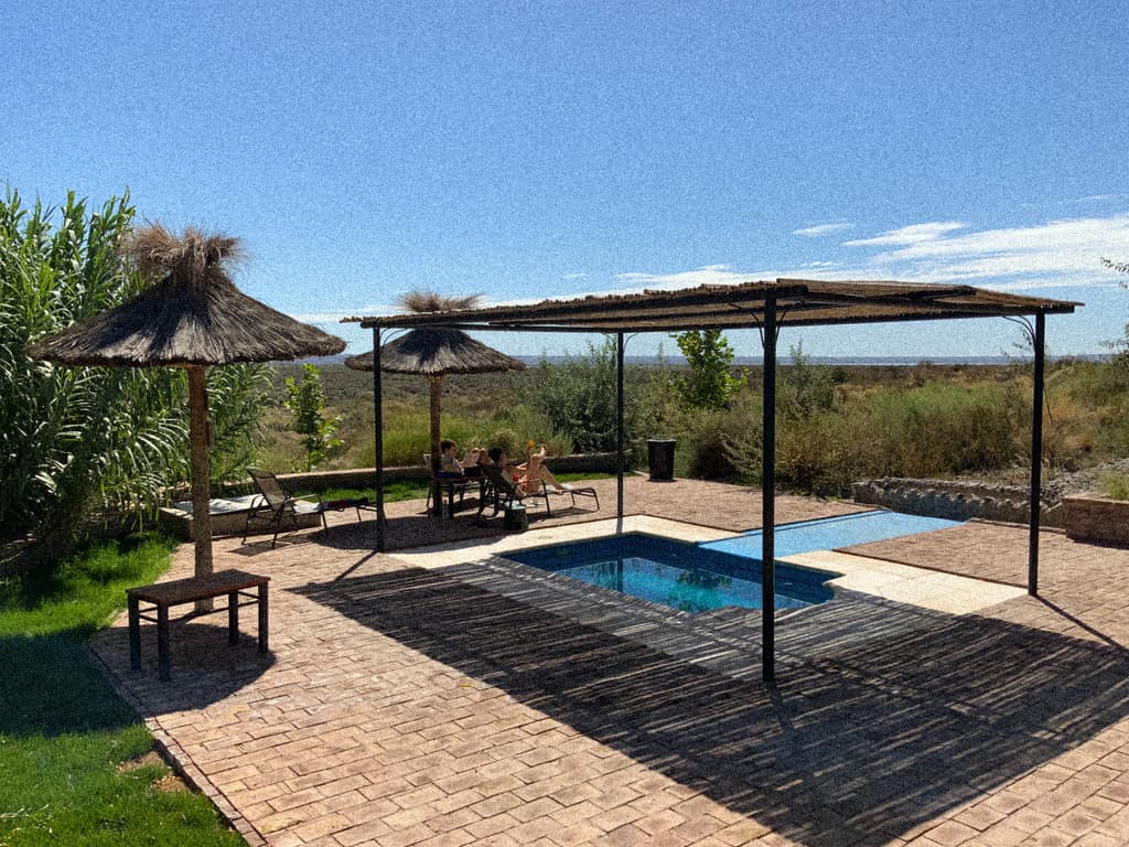 La pileta de la terraza bajo una pérgola de caña, con dos sombrillas de paja, gente tomando sol y la meseta detrás.