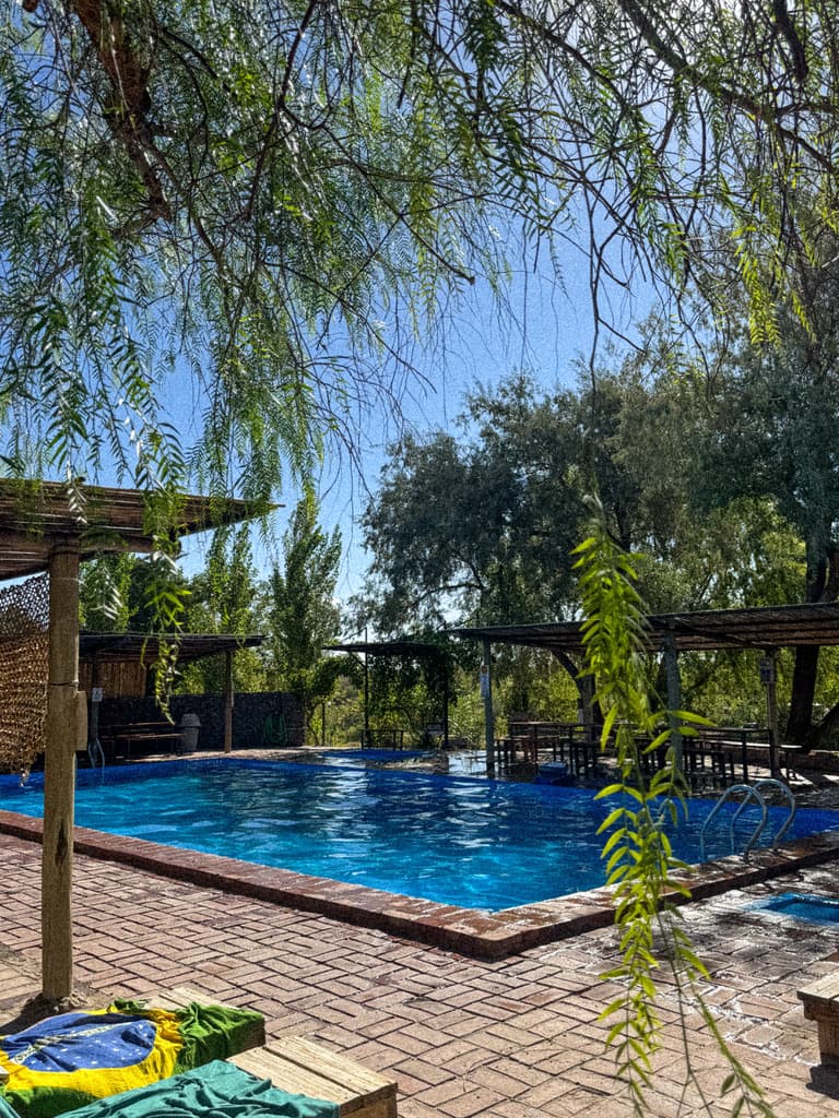 La pileta grande vista desde abajo de un árbol, con las mesas bajo los techos de caña al fondo y dos toallas sobre un banco.