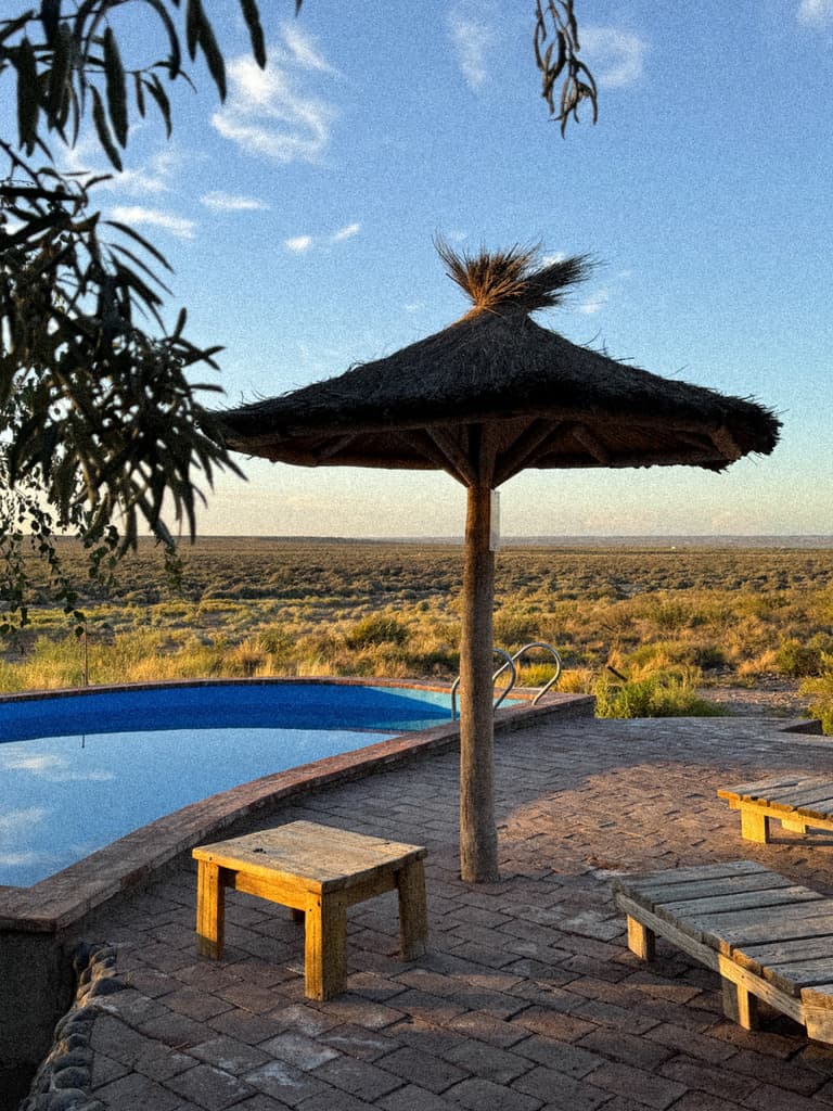 Una sombrilla de paja junto a la pileta media luna, con reposeras de madera y la meseta al fondo.